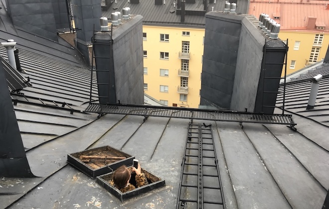 Näkymässä kiinteistön katot ja ihminen katsoo kattoikkunasta ja ottaa valokuvan näkymästä.